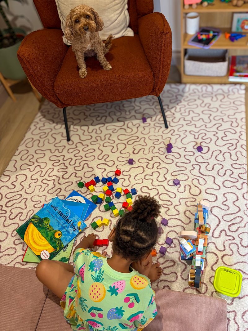 babs watching from the armchair while a child plays with toys on the rug